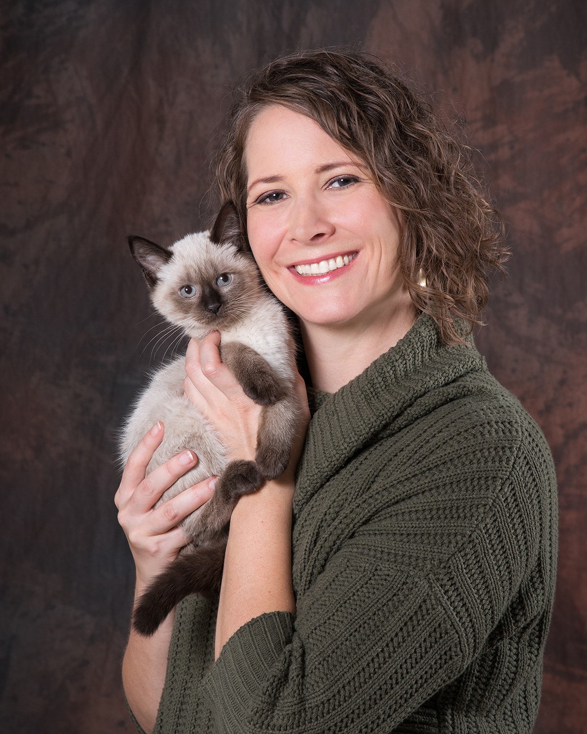 Heather Miller smiling in a green sweater. She is holding a siamese mix kitten up near her face.