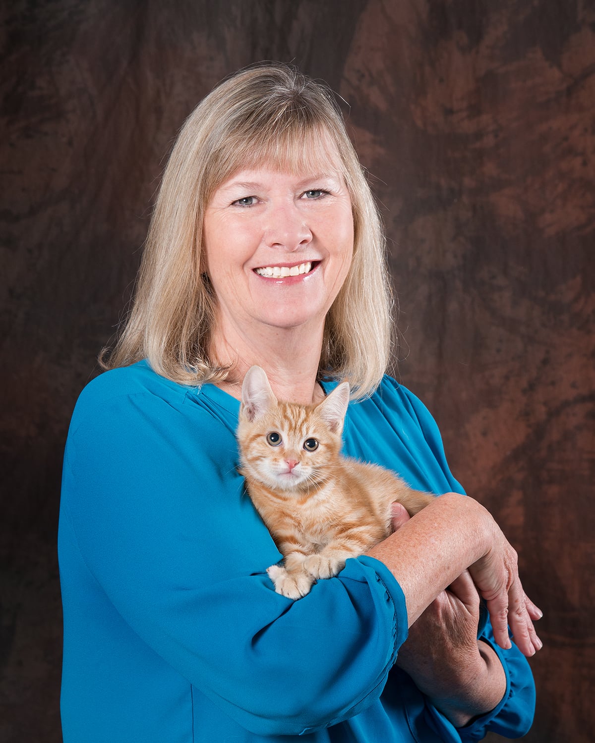 A headshot of Nancy Puro. She is smiling in a blue blouse and holding an orange kitten.