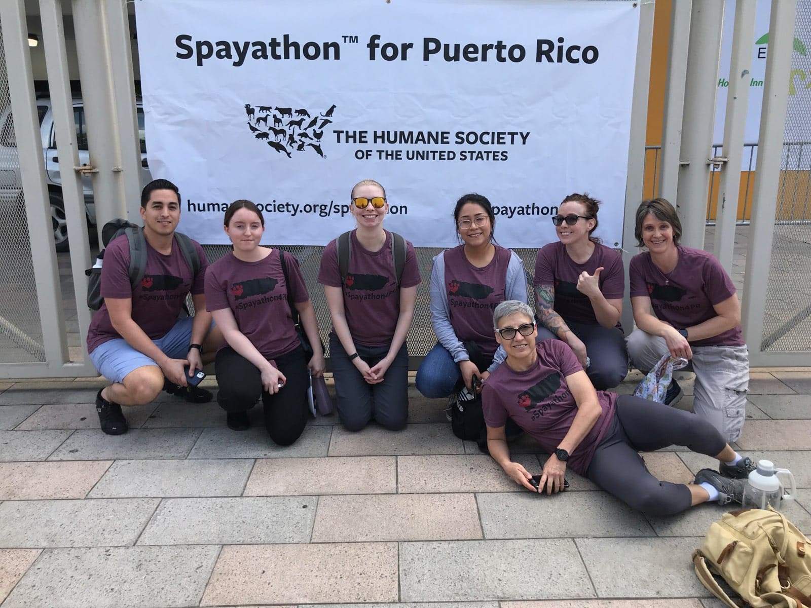 Six people kneel and one person lounges on the ground in front of a white banner advertising the Spayathon for Puerto Rico.
