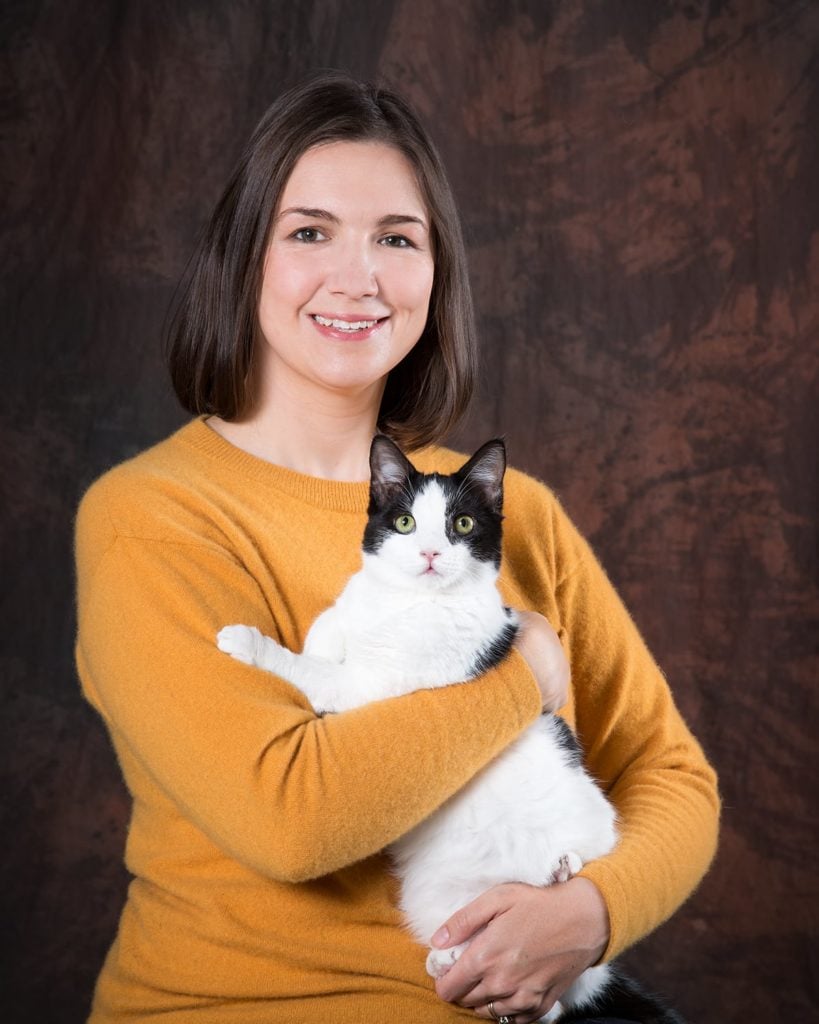 A headshot of Brittany Perkins holding a black-and-white cat. She is wearing a yellow sweater.