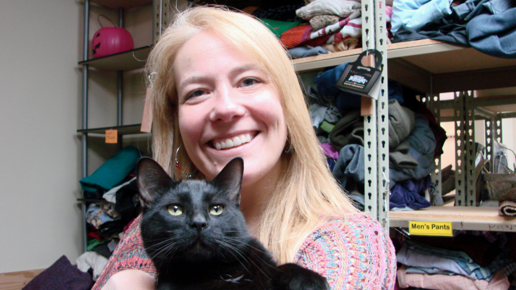 Robin Eckstein, a volunteer at CAT's thrift store, holds a black cat in her arms while standing in front of a shelf full of donated items.