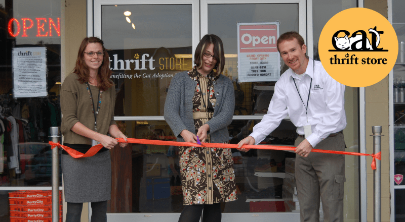 At the CAT Thrift Store Ribbon Cutting Ceremony, two CAT staff members smile as they hold a red ribbon across the store's front doors while a third staffer cuts the ribbon with scissors.