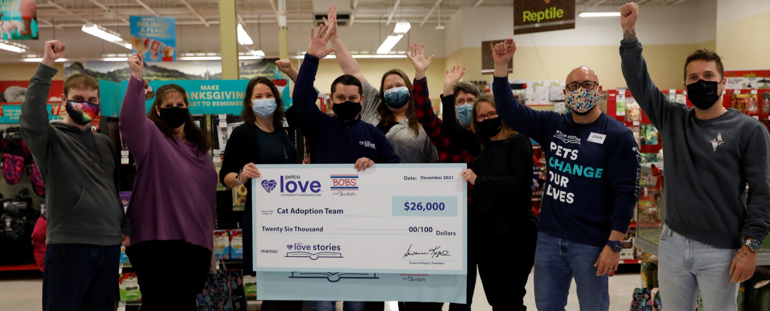 A group of people in face masks raise their hands in celebration behind a large novelty check for $26,000 to Cat Adoption Team from Petco Love.