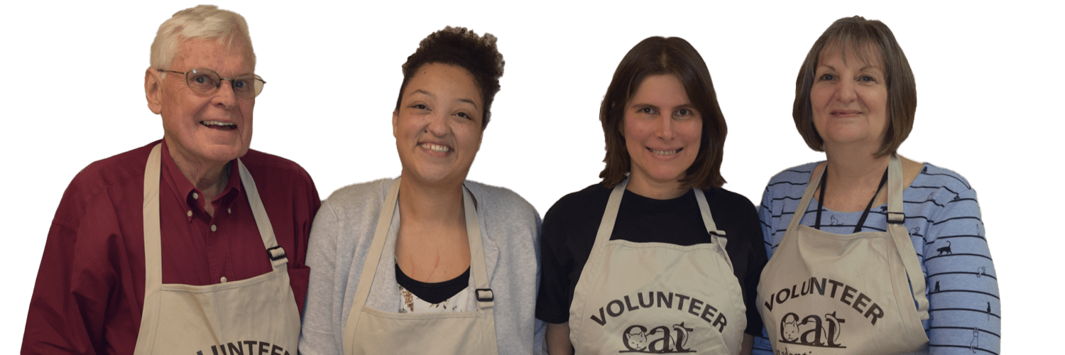 Four smiling volunteers of different ages, ethnicity, and genders stand shoulder-to-shoulder wearing their tan CAT Volunteer aprons over their street clothes.