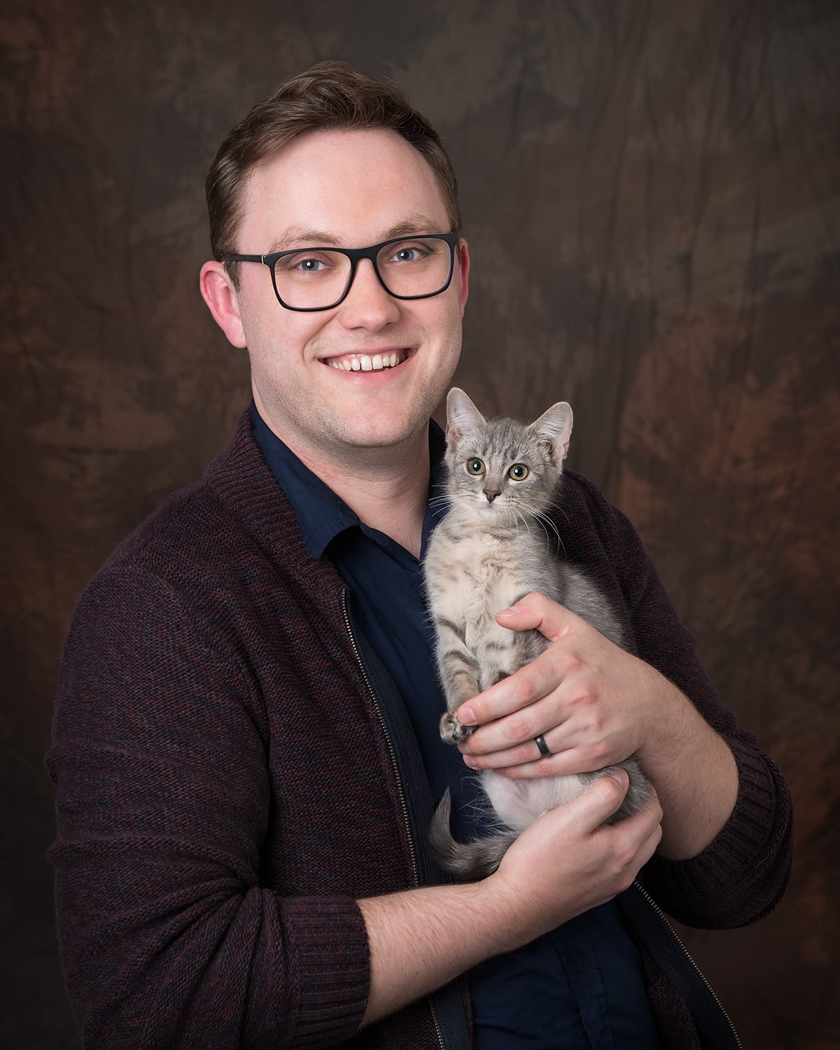 Headshot of Josh Kneeland in a blue button-up shirt and dark-colored cardigan. He wears glasses and is holding a gray kitten.