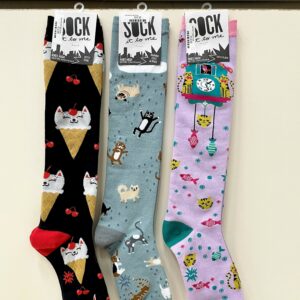 Three knee-length socks are shown hanging in a row. The left sock is black with ice cream cones and cat faces as the "scoop" of ice cream. The middle sock is blue with cats and dogs "raining" down along with rain drops. The sock on the right is light purple with a cuckoo clock, tabby cats and fish decorating it.