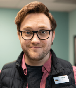 Headshot of Josh in a closed-mouth smile, wearing dark-rimmed glasses, a collared shirt, a black vest and his nametag.