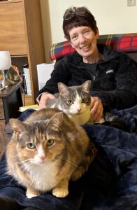 Sue sits, leg extended, on a chair. Her cats Cammie and Hunter snuggle the length of her legs on a blanket.