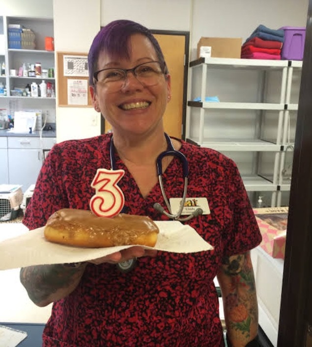CAT staff member in scrubs holds up a donut with a 3 candle stuck in it