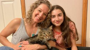 A mother and daughter sit close together with a longhaired tabby cat sitting on their laps.