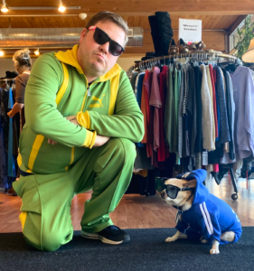 Hansel, in green-and-yellow track suit, kneels and strikes an arms-crossed pose. Next to him, in a blue tracksuit, is Franky, a small, beige dog in a blue tracksuit. They both wear sunglasses.