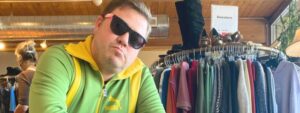 Hansel, wearing a green and yellow track suit and sunglasses, in front of clothing racks inside CAT Thrift Store.