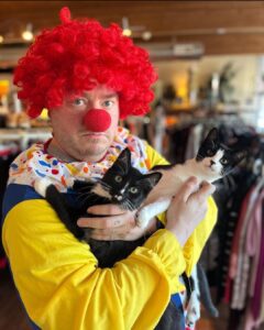 Hansel wears a clown suit, with a red wig and nose. He holds two kittens, Bo and Blarry, prior to their adoption.