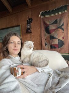 Mara and Sticky Rice snuggle on a comfy chair. The background is wood paneling. Mara has a mug in their left hand. Sticky looks past the frame of the photo to what we assume is out the window.