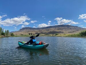 Lake, mountains, and blue sky with a few white clouds fill the frame. A person in a hat and blue personal flotation device rows away from us in a kayak.