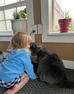 Hilda sits on the floor next to a young child in a blue top. They touch noses. Photo taken from behind. The two are in profile.