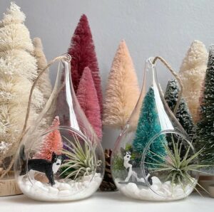 Colorful bottlebrush Christmas trees behind two small glass terrariums filled with cat figurines, fake snow, and an air plant.