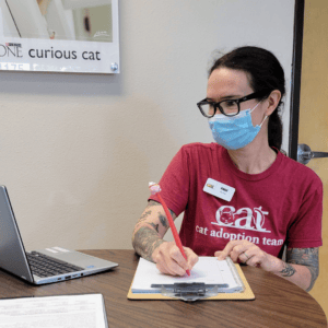 Anna, in a red CAT shirt and nametag, sits at a table, wearing a face mask. She writes on a clipboard on the table in front of her.