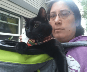 Carrie and Hossa, black cat, stand outside a home. Hossa is in a green-and-grey cat pack on Carrie's front. Straps are seen.