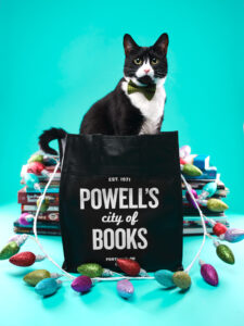 A tuxedo cat sits on top of a bile of books behind a black Powell's Books reusable shopping bag.