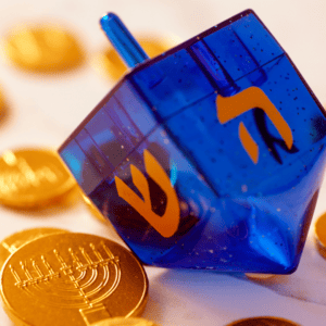Blue toy dreidel sits on its side with two Hebrew letters in gold showing. Background is Hanukkah gold-wrapped foil coins on the white table.