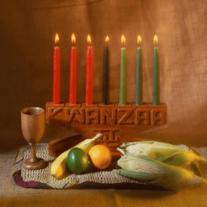 Wooden candle holder that says, "Kwanzaa" on the center of it. It holds seven candles in red, black, and green. It sits on a table with a few symbolic food items.