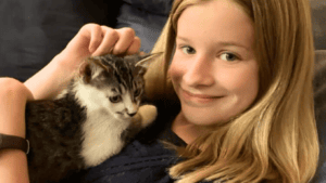 Closeup of a child with blonde hair smiling into the camera with a brown-and-white kitten on their chest.