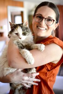 A person in glasses and an orange shirt holds Schmutz in their arms. Schmutz is mostly white with some grey patches on her face and paws.