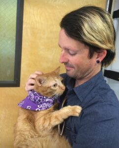 Garrett holds Marmalade and orange cat wearing a purple bandana around his neck.