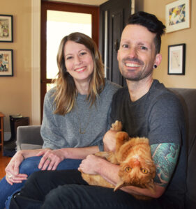 Two people sit side-by-side on a couch smiling into the camera. The person on the right holds an upside-down orange cat.