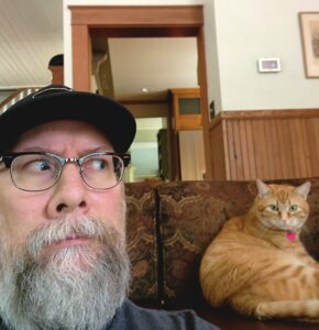 A bearded person in close up sits next to an orange cat on a couch. They look at one another