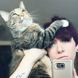 A striped cat climbs a person's head. The person holds a phone and looks into the lens.