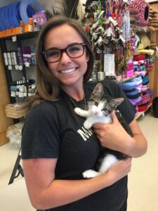 A person in a black tee shirt and glasses smiles while holding a small brown-and-white kitten.