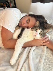 A person in a white top and a brown-and-white cat snuggle on a bed. The person's eyes are closed and they cat's are open.