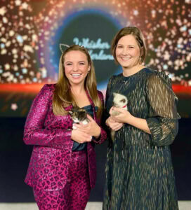 Two people stand in front of a screen, wearing cat ears and holding kittens. They are dressed for a classy event.